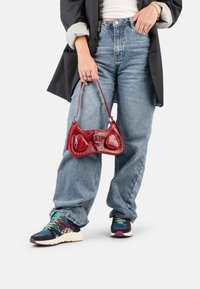Una persona indossa un blazer nero sopra una maglietta bianca e jeans larghi blu, mentre tiene una piccola borsa rossa, completata da scarpe da ginnastica colorate.