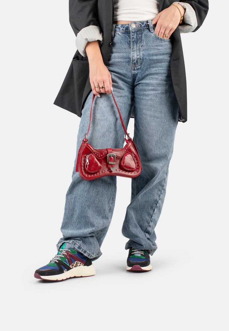 Una persona indossa un blazer nero sopra una maglietta bianca e jeans larghi blu, mentre tiene una piccola borsa rossa, completata da scarpe da ginnastica colorate.