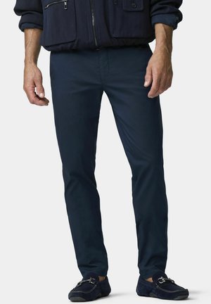 Mann trägt marineblaue, schlank geschnittene Hose, marineblaue Jacke mit Reißverschluss und Taschen sowie marineblaue Loafers mit Metallbeschlag.