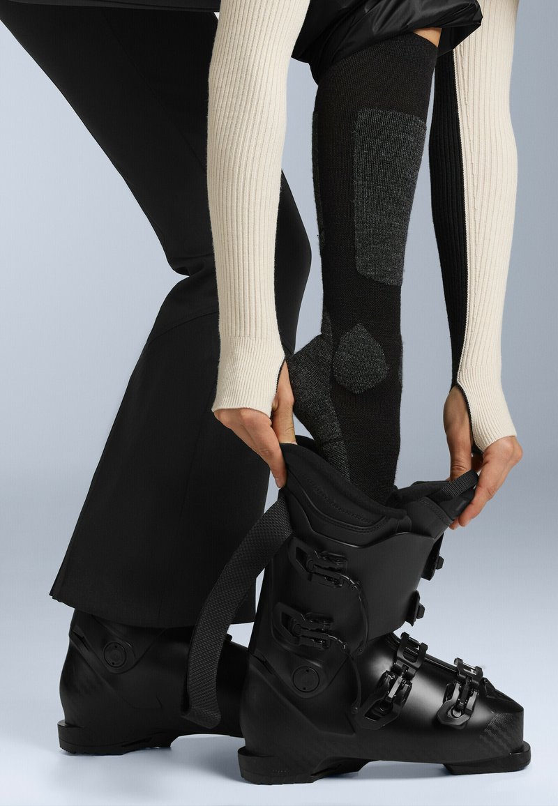 Svart skidsko med glänsande yta och fyra justerbara spännen, som lyfts av händer. Ribbad ljusfärgad ärm står i kontrast till mörka knästrumpor.