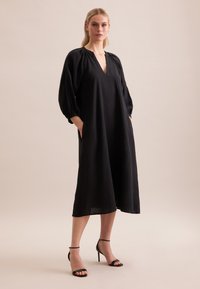 Abito lungo nero con scollatura a V e maniche gather. Realizzato in tessuto leggero, presenta tasche laterali e una vestibilità comoda. Tacchi neri con cinturini.