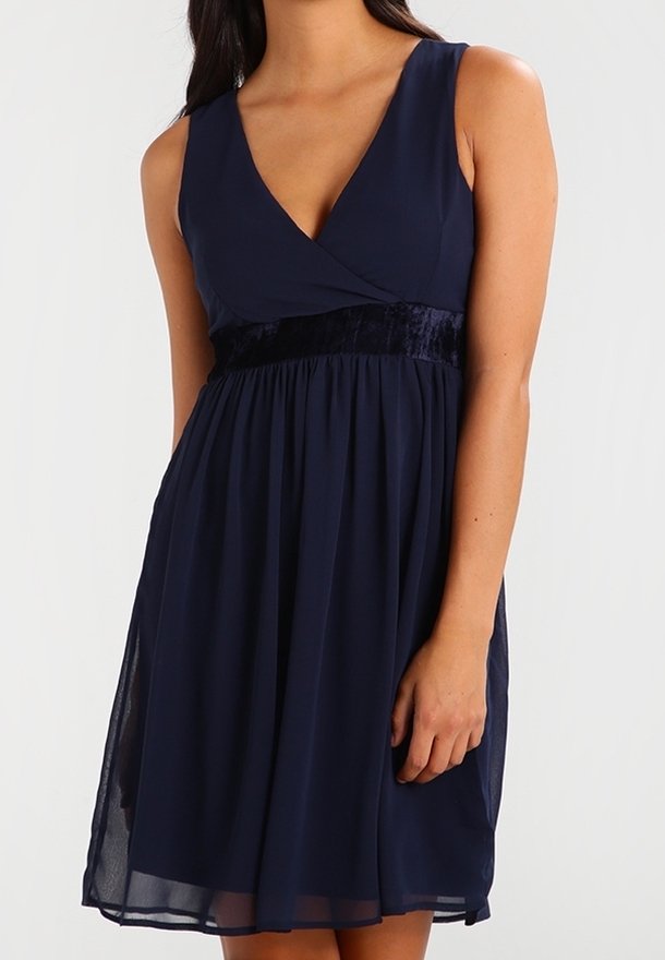 Robe bleu marine sans manches avec col en V et ceinture en velours, jupe fluide tombant au-dessus des genoux, portée par une femme debout.