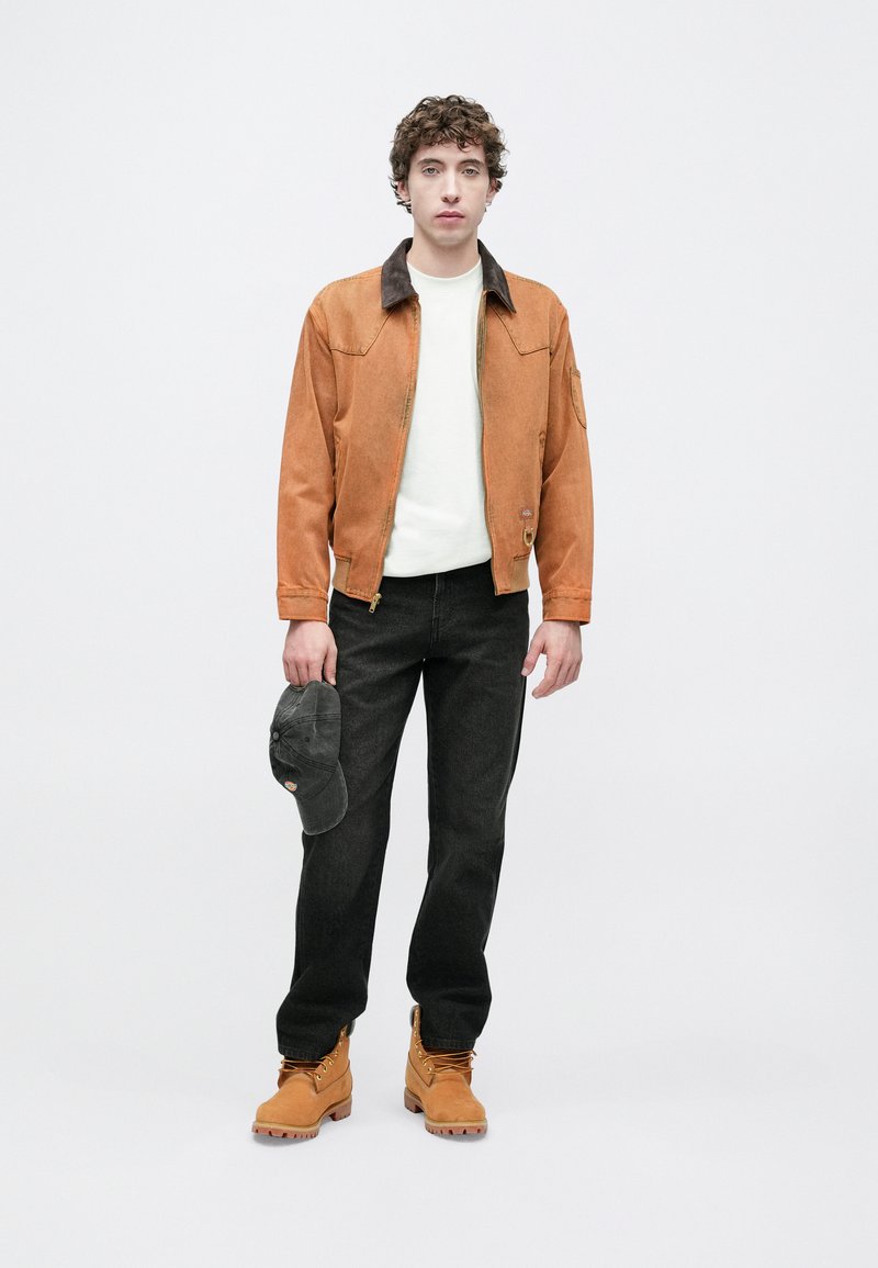 Light brown suede jacket with a dark collar, white sweater, black jeans, and tan boots. Holding a gray cap, standing against a white background.