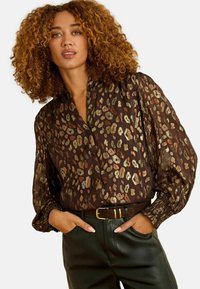 Blusa trasparente marrone con stampa leopardata metallica, dotata di scollatura a V, maniche lunghe a sbuffo e polsini arricciati. Abbinata a una cintura e pantaloni in pelle.