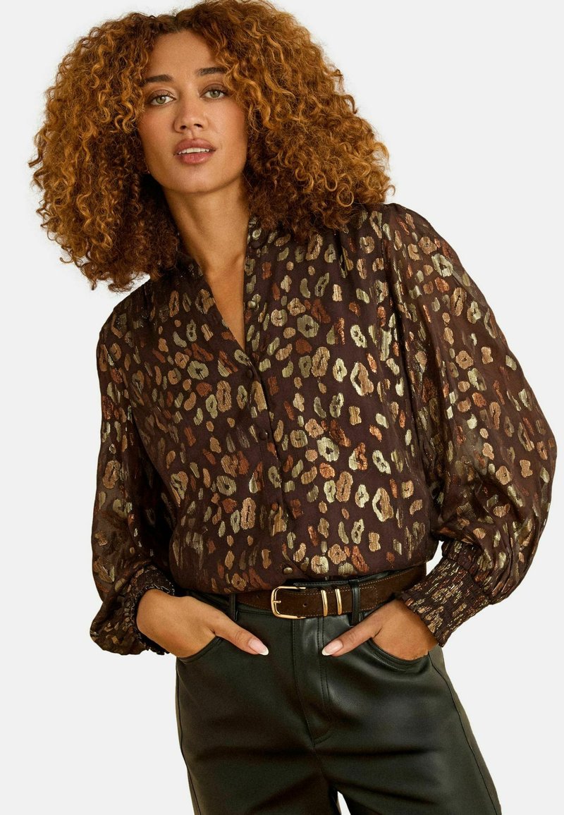 Blusa trasparente marrone con stampa leopardata metallica, dotata di scollatura a V, maniche lunghe a sbuffo e polsini arricciati. Abbinata a una cintura e pantaloni in pelle.
