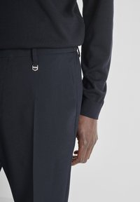 Marineblauwe broek van gladde stof, met een accent van zilveren hardware en een getailleerde pasvorm, gecombineerd met een donkere trui.