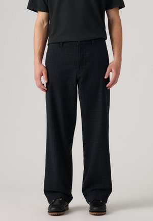 Hombre vestido con pantalones negros sueltos, camiseta negra y zapatos negros con suelas marrones, de pie contra un fondo claro y simple.
