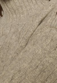 Närbild av en beige kabelstickad ylletröja med en ribbad polokrage, bärs av en person.