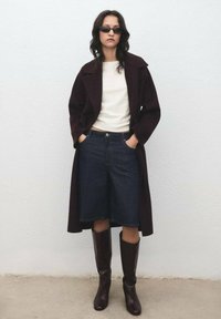 Bordový kabát cez bielu košeľu, doplnený tmavomodrými denimovými culottes a hnedými vysokými čižmami; model stojí s rukami v vreckách pri svetlej stene.
