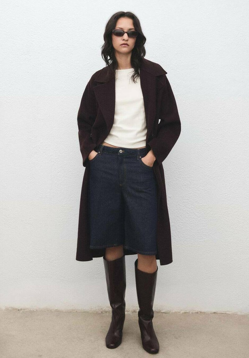 Bordový kabát cez bielu košeľu, doplnený tmavomodrými denimovými culottes a hnedými vysokými čižmami; model stojí s rukami v vreckách pri svetlej stene.