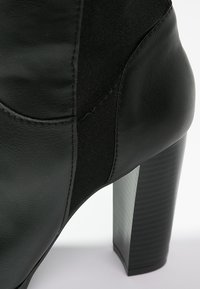 Stivale alla caviglia nero in pelle e suede con tacco alto, che mostra dettagli del tacco e cuciture laterali su uno sfondo bianco.