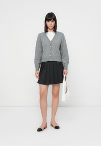 Grau gestrickte Strickjacke mit Knöpfen, kombiniert mit einem schwarzen Faltenrock und flachen schwarzen Schuhen. Hält eine weiße Handtasche. Schlichtes Design und Texturen.