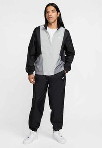 Φόρμα της Nike με γκρι και μαύρο τζάκετ με φερμουάρ και ελαστικά μανίκια, σε συνδυασμό με μαύρο παντελόνι και λευκά αθλητικά παπούτσια.