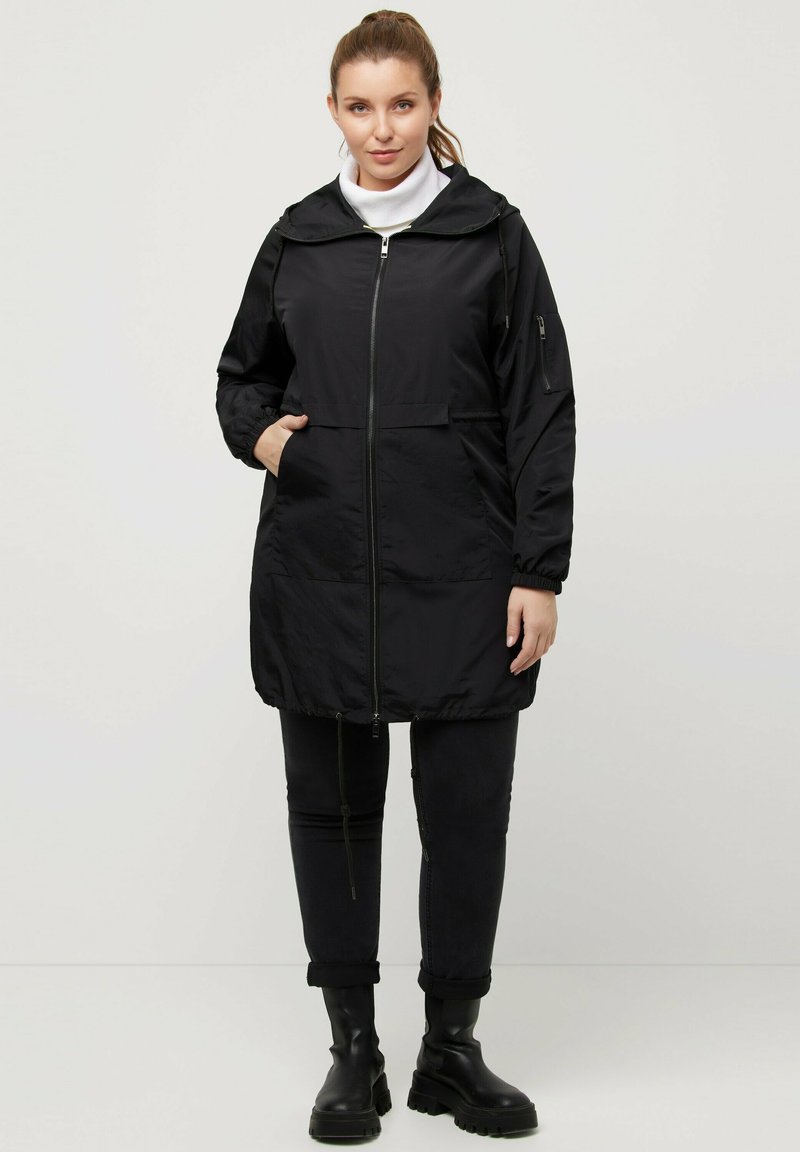 Svart vindjacka med dragkedja fram, huva, elastiska ärmslut och sidofickor. Tillverkad av ett tunt, lätt material.