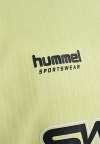 Lys gul sportswear-stof med en tekstureret overflade, prydet med et sort "hummel" logo og "SPORTSWEAR" tekst i fed skrift.