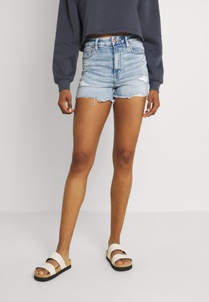 Personne portant un short en denim déchiré bleu clair, un sweat-shirt court à manches longues foncé, et des sandales blanches avec des semelles en liège.