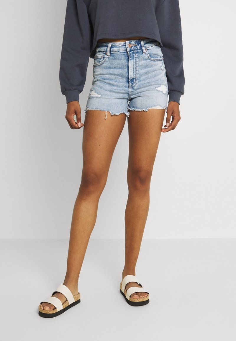 Personne portant un short en denim déchiré bleu clair, un sweat-shirt court à manches longues foncé, et des sandales blanches avec des semelles en liège.