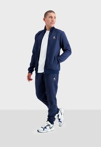 Tuta da allenamento blu navy realizzata in tessuto liscio, con giacca con zip e logo e pantaloni abbinati. Indossato con scarpe da ginnastica bianche e blu navy.
