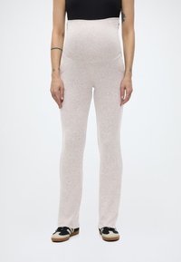 Γυναίκα που φοράει ανοιχτό μπεζ παντελόνι εγκυμοσύνης με ψηλή μέση, μαύρο crop top και μαύρα-άσπρα αθλητικά παπούτσια, σκιασμένη μπροστά σε απλό λευκό φόντο.