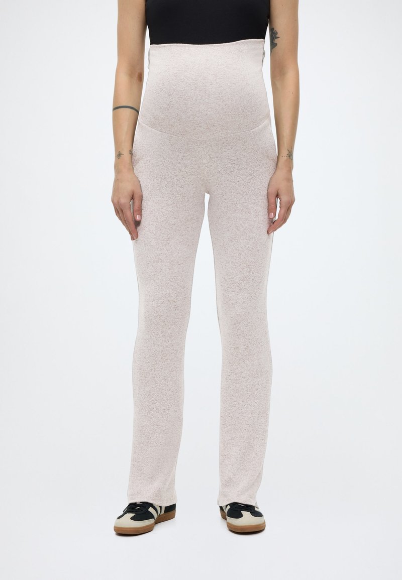 Γυναίκα που φοράει ανοιχτό μπεζ παντελόνι εγκυμοσύνης με ψηλή μέση, μαύρο crop top και μαύρα-άσπρα αθλητικά παπούτσια, σκιασμένη μπροστά σε απλό λευκό φόντο.