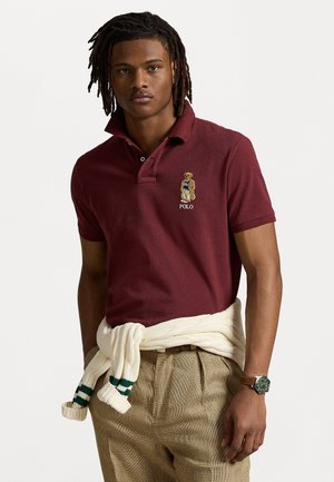 T-shirt polo bordeaux avec logo d'ours brodé et texte "Polo". Porté avec un pantalon à carreaux beige et un pull blanc noué à la taille.