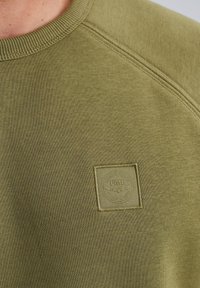 Sweatshirt vert olive en tissu texturé avec un col rond, des manches raglan et un patch logo discret sur la poitrine.
