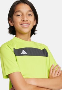 Camiseta deportiva de color verde neón con una franja horizontal negra y un logotipo blanco. Mangas cortas, cuello redondo y tejido suave y ligero.