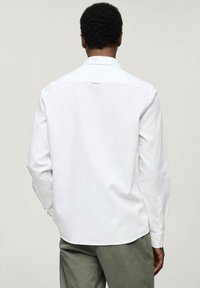 Camisa blanca de manga larga con cuello clásico, cierre de botones y un detalle de lazo en la parte posterior, combinada con pantalones verde oliva.