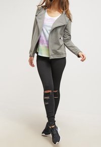 Donna che indossa una felpa grigia con zip, un top a canotta multicolore con fantasia, jeans skinny neri strappati e sneaker nere, mentre cammina in avanti.