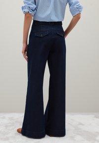 Pantaloni in denim blu scuro a gamba larga con due tasche posteriori, chiusure a bottone e orlo pulito. Abbinati a una camicia a righe blu e bianche.