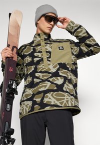 Fleece pulóver fekete és olajzöld camo mintával, félgombos záródással, mellzsebbel, szürke sapkával és sífelszereléssel párosítva.