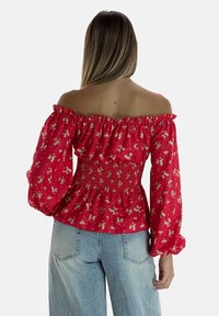 Blusa rossa floreale off-shoulder con maniche a sbuffo, vita elasticizzata e un motivo testurizzato. Abbinata a jeans in denim azzurro chiaro.