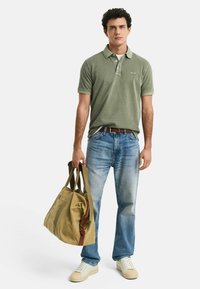 Grünes Poloshirt mit Kragen, blaue Jeans, beigen Sneakers und eine sandfarbene Reisetasche mit braunen Akzenten, gehalten von einem stehenden männlichen Modell.