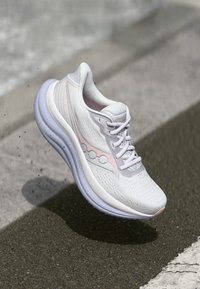 Chaussure de sport blanche avec un dessus texturé, des accents roses et une semelle épaisse et rembourrée, présentée en suspension au-dessus d'une surface sombre.