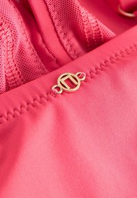 Soutien-gorge rose en tissu lisse, orné de dentelle et d'une boucle logo en ton or. Comprend des bretelles réglables et des coutures le long des bords.