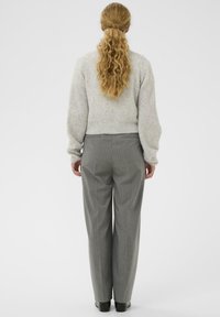 Asmuo su ilgais banguotais plaukais, surištais į uodegą, dėvi šviesiai pilką megztinį ir pilkai dryžuotas kelnes, stovėdamas prieš paprastą baltą foną.