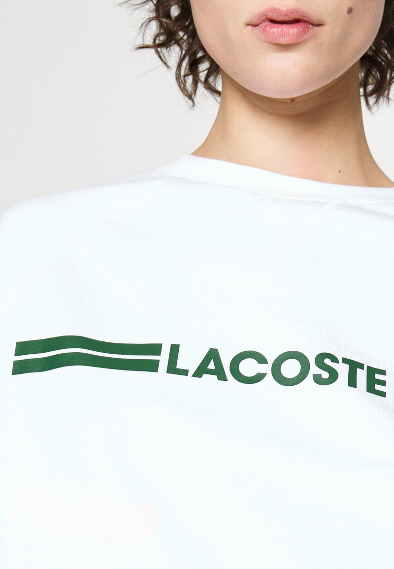 Sweat-shirt blanc en coton, arborant un logo "LACOSTE" vert audacieux avec deux lignes horizontales. Tissu texturé et col côtelé.