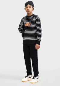 Pulover cu dungi alb-negru, pantaloni negri, pantofi Nike albi cu galben și geantă neagră purtată transversal, completate de o căciulă neagră.