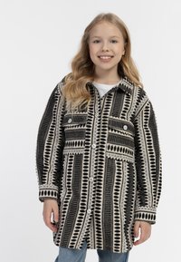 Ylin roikkuva musta ja kerma kuviollinen takki, jossa on suuret napit, kaksi etutaskua ja kauluksellinen kaulus. Teksturoitu kangas, jossa on vaihteleva kuvio.