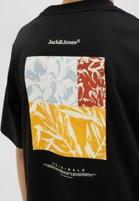 Tricou negru cu un grafic pătrat multicolor în galben, roșu și albastru, cu modele abstracte; numele brandului imprimat deasupra designului.