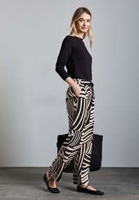Black long-sleeve top paired with high-waisted beige pants featuring bold black stripes; model holds a black bag, wearing flat shoes.