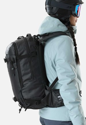 Person wearing black ski helmet, goggles, light blue jacket, and large black backpack with multiple straps and buckles, facing right.