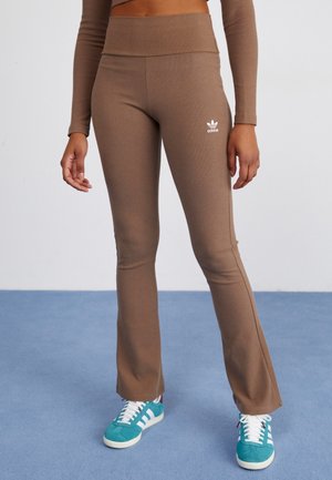 Kobieta ubrana w brązowe rozszerzane legginsy Adidas i turkusowe buty sportowe Adidas stoi na niebieskim dywanie na tle białej ściany.