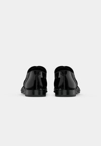 Un paio di scarpe formali in pelle nera lucida viste dal retro su uno sfondo grigio chiaro uniforme.