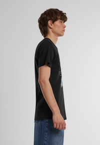 Melnas kokvilnas T-krekls ar īsām piedurknēm, kuram priekšpusē ir grafiskais dizains. Griezums ir regulārs, un audums izskatās gluds.