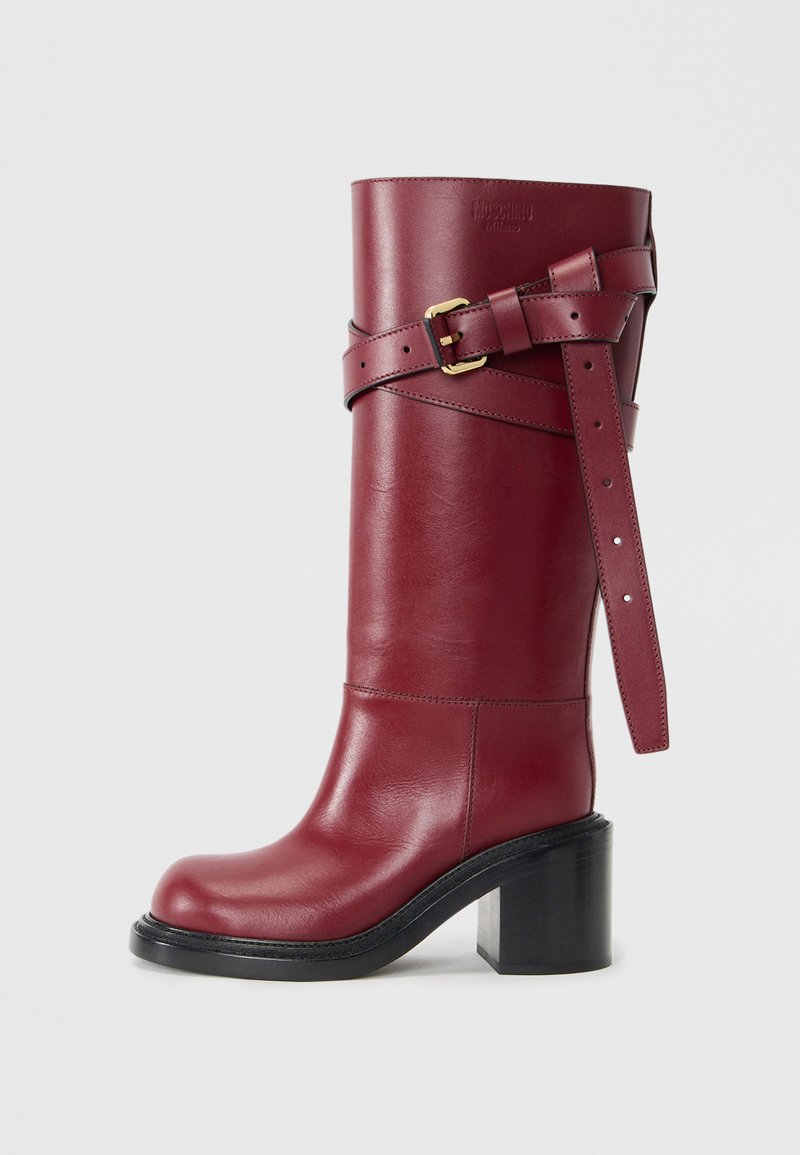 Red leather knee-high boot with a chunky black heel, features a wide strap with a gold buckle and adjustable holes, smooth texture.