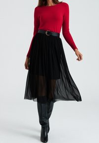 Röd åtsittande tröja i kombination med en svart plisserad maxikjol, accentuerad av ett svart bälte med en rund guldfärgad spänne och knähöga svarta stövlar.