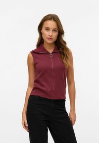 Vinröd ärmlös topp med randen krage, försedd med en fron dragkedja och ett runt metallhänge. Kombinerad med svarta byxor.