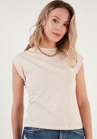 Top in cotone beige con maniche corte arrotolate e scollatura rotonda. Indossato con jeans blu, presenta una texture liscia e una silhouette aderente.