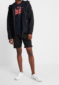 Chaqueta negra con cremallera y acentos azules, camiseta oscura con un gráfico rojo, pantalones cortos negros y zapatillas blancas. Texturas suaves en todo.